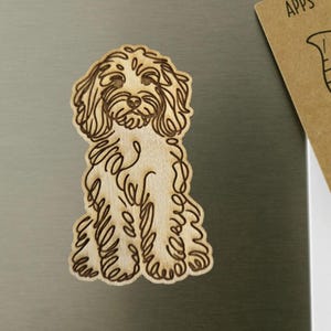 May include: A wooden cutout of a dog, featuring a detailed line drawing. The dog is light brown with darker brown etched lines, sitting upright. The cutout is on a metallic surface, with a small brown card in the background.