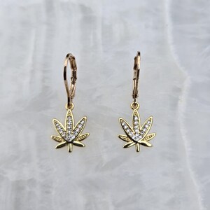 May include: A pair of gold-colored earrings featuring a cannabis leaf design. Each leaf is embellished with clear, sparkling gemstones. The earrings have a lever-back closure and are displayed against a light, marbled background.