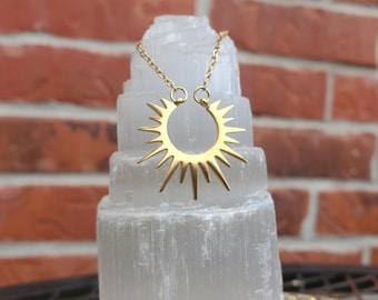 May include: A gold-colored sunburst pendant necklace. The pendant is a stylized sun with pointed rays. The necklace is displayed on a white, layered stone structure against a brick wall background.