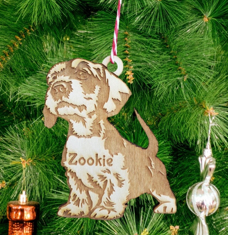 May include: A wooden Christmas ornament shaped like a dog with the name "Zookie" engraved on its chest. The ornament is hanging on a Christmas tree.