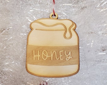 Pode incluir: Enfeite de madeira em forma de pote de mel com a palavra "HONEY" gravada na frente. O enfeite de madeira clara pende de um fio vermelho e branco. O fundo é uma superfície branca e brilhante.