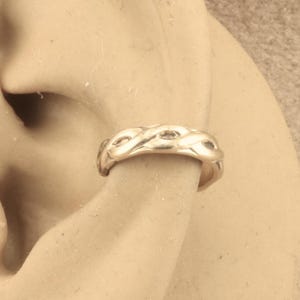 May include: A gold-colored ear cuff with a woven design. The cuff is worn on the ear and has a continuous, braided pattern. The jewelry is made of a shiny, metallic material.