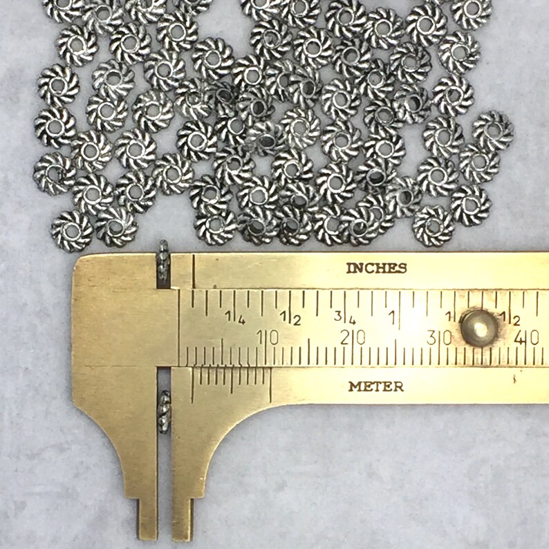 May include: A close-up of a ruler measuring a pile of silver metal beads. The beads are about 1/2 inch in diameter and have a swirl design.