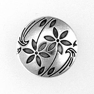 May include: A round, silver-toned button with a floral design. Two stylized flowers with dark centers and petals are featured, along with curved leaf and stem accents. The button is set against a white background.