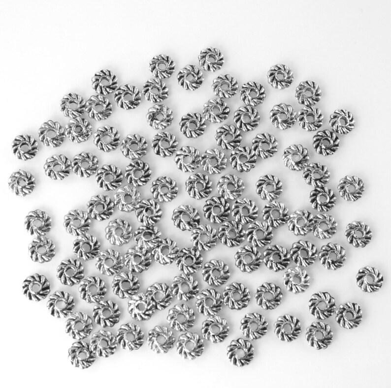 May include: A large quantity of silver-toned metal beads with a twisted design. The beads are scattered on a white surface.
