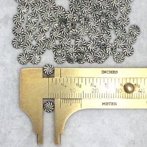 May include: A pile of silver metal spacer beads with a twisted design. The beads are approximately 1/4 inch in diameter. A ruler is used to show the size of the beads.