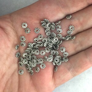 May include: A pile of silver metal beads with a decorative swirl design. The beads are small and round, and they are perfect for jewelry making.