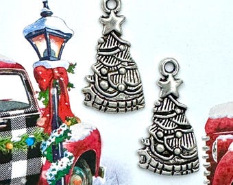 May include: Three silver-colored Christmas tree charms with star toppers. The charms are detailed with etched lines and circular ornaments. The background features a red truck with a Christmas wreath and a snow-covered lamp post.