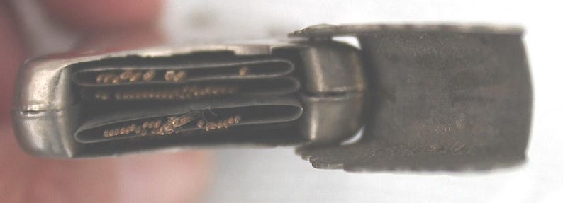 May include: A small, antique silver-toned metal case, partially open, revealing folded paper inserts. The case has a rectangular shape with rounded edges. The paper inserts have text and are folded in a concertina style.