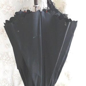 Op de afbeelding: Een vintage zwarte paraplu met een gebogen, sierlijke handgreep. De handgreep is donkergrijs en de paraplu heeft een houten handvat. De stof van de paraplu is zwart en heeft een geschulpte rand. Een zwarte kwast hangt aan de handgreep.