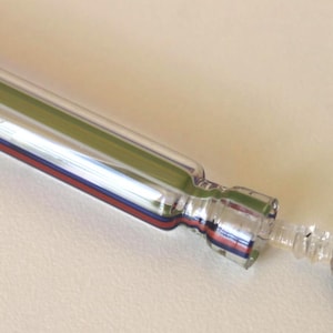 Puede incluir: Un tapón de botella de vino de vidrio transparente con un cuerpo largo y cilíndrico. El tapón tiene una franja verde en el centro, con rayas rojas y azules a cada lado. El extremo tiene un sello de goma negro y una punta transparente texturizada.