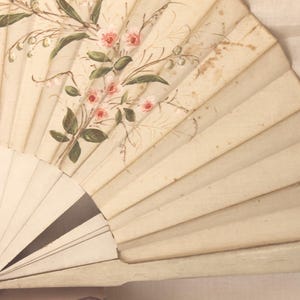 Pode incluir: Um leque de mão vintage com fundo creme. O leque apresenta um design floral com rosas cor-de-rosa e folhas verdes. O leque está parcialmente aberto, revelando sua estrutura plissada.