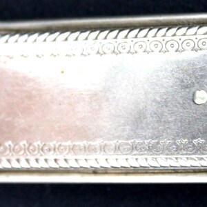 May include: A rectangular, silver-toned antique case with ornate detailing. The case features a decorative rope-like border and a central panel with a lighter, possibly copper-toned, section. A small, circular clasp is visible on the right side.