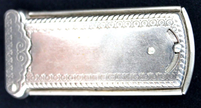 May include: A rectangular, silver-toned antique case with ornate detailing. The case features a decorative rope-like border and a central panel with a lighter, possibly copper-toned, section. A small, circular clasp is visible on the right side.