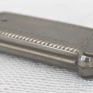 May include: A silver-toned, rectangular antique cigarette case with ornate detailing. The case features a decorative rope-like border and the word "REDDITCH" is visible on the end. The case is closed and appears to be made of metal.