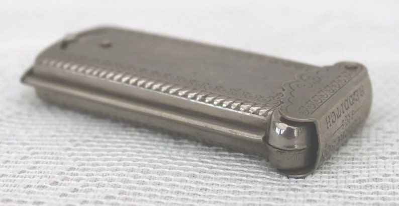 May include: A silver-toned, rectangular antique cigarette case with ornate detailing. The case features a decorative rope-like border and the word "REDDITCH" is visible on the end. The case is closed and appears to be made of metal.