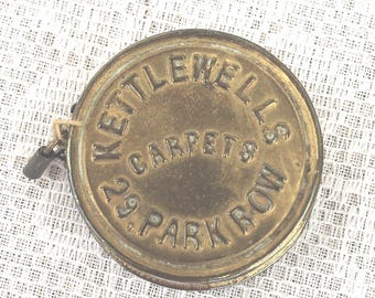May include: A vintage brass tape measure with the text "Kettlewell's Carpets 29 Park Row" embossed on the circular casing. The tape measure has a small metal loop and a clasp attached. The casing has a tarnished, aged appearance.