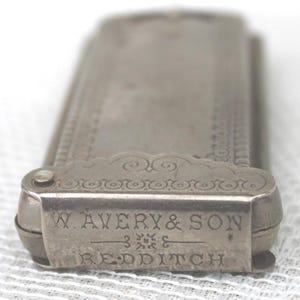 May include: Close-up of a silver metal object with the text "W. AVERY & SON REDDITCH" engraved on the front. The object has a rectangular shape with decorative scrollwork and a hinged lid. The object appears to be a vintage item.
