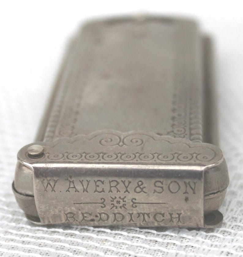 May include: Close-up of a silver metal object with the text "W. AVERY & SON REDDITCH" engraved on the front. The object has a rectangular shape with decorative scrollwork and a hinged lid. The object appears to be a vintage item.