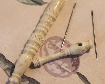 May include: A cream-colored, carved needle case with a screw-top lid and a matching smaller component featuring a single hole, accompanied by two thin, metallic sewing needles. The needle case has a textured, geometric pattern along its length.