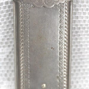 May include: A rectangular, silver-toned antique case with ornate detailing. The case features a hinged lid with decorative scrollwork and a textured border. The body has a central panel with a small, curved indentation and two small, round accents.