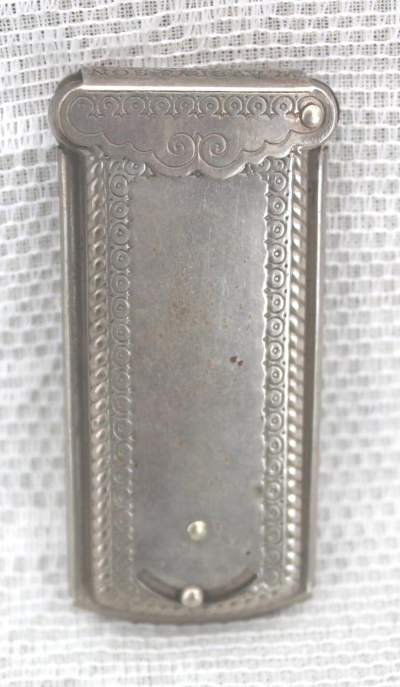 May include: A rectangular, silver-toned antique case with ornate detailing. The case features a hinged lid with decorative scrollwork and a textured border. The body has a central panel with a small, curved indentation and two small, round accents.