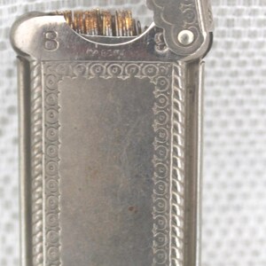May include: A vintage silver-toned lighter with an ornate design. The lighter has a rectangular body with decorative borders and a hinged lid. The lid is open, revealing the wick and flint. The number "8" is visible.
