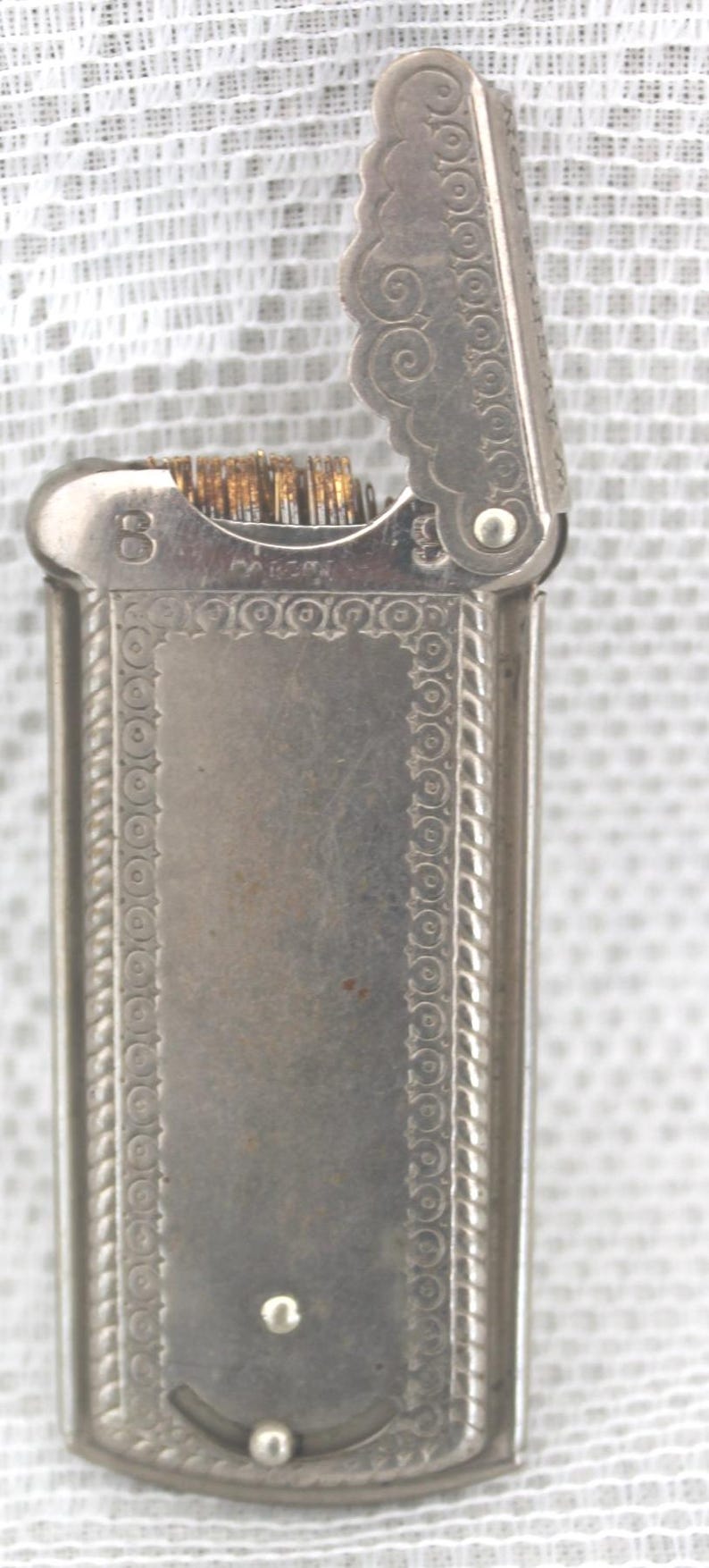 May include: A vintage silver-toned lighter with an ornate design. The lighter has a rectangular body with decorative borders and a hinged lid. The lid is open, revealing the wick and flint. The number "8" is visible.