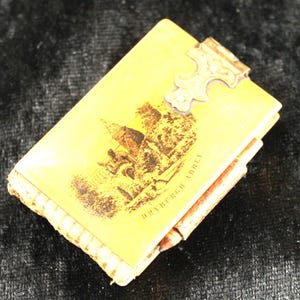 Puede incluir: Un pequeño libro antiguo rectangular con una cubierta amarilla y un broche de metal. La cubierta presenta una ilustración en tonos sepia de un edificio y el texto "DRYBURGH ABBEY". Los bordes del libro están desgastados, lo que sugiere antigüedad.