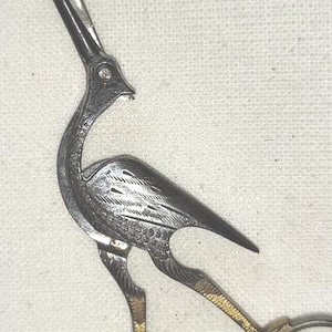 May include: Vintage metal scissors in the shape of a bird, likely a crane or heron. The scissors have a detailed design, with etched feathers and a long, pointed beak. The handles are gold-toned, and the blades are silver-toned.