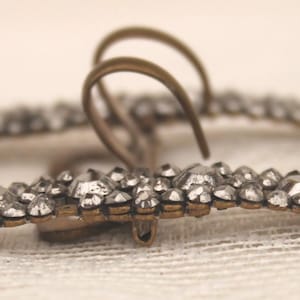 May include: A vintage silver-coloured buckle, rectangular in form, adorned with many small, glittering stones. It has two arched loops and a small clasp, likely for securing clothing or accessories. The design is intricate and detailed, with a classic aesthetic.