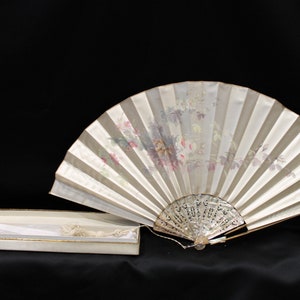 May include: A cream-coloured hand fan with a floral pattern and a decorative handle. The fan is next to a matching box with a tassel. The fan's design includes pink, purple, and green floral elements.