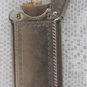 May include: Antique silver-toned match safe with an open lid. The rectangular case features ornate detailing, including a decorative border and a hinged lid. The number "8" is visible on the top. The match safe contains several matches.