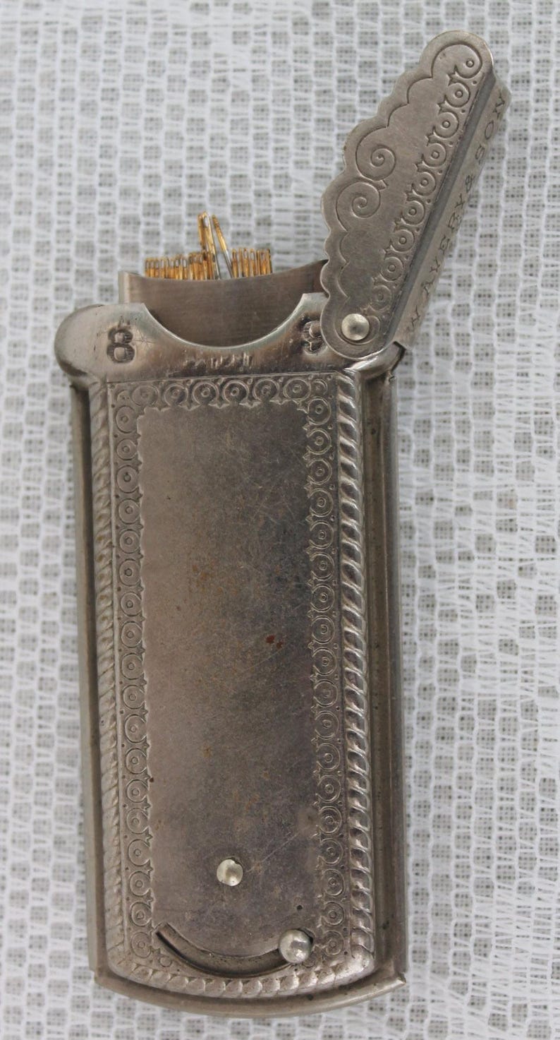 May include: Antique silver-toned match safe with an open lid. The rectangular case features ornate detailing, including a decorative border and a hinged lid. The number "8" is visible on the top. The match safe contains several matches.