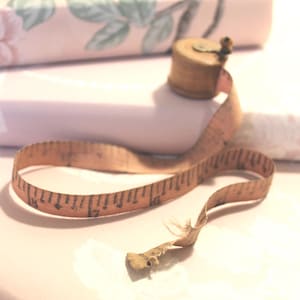 May include: A vintage wooden tape measure with a retractable pink measuring tape, resting on a stack of floral patterned paper rolls. The tape measure is brown and the measuring tape is marked with black numbers.