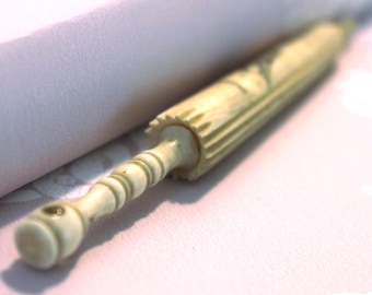 May include: An antique ivory-colored rolling pin with a detailed, carved design. The rolling pin has a cylindrical body with a ridged section and a decorative handle. The handle has a rounded end with a small hole. The rolling pin is resting on a light pink surface.