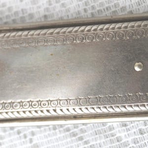 May include: A rectangular, silver-toned metal object with ornate detailing. The piece features a decorative border with a rope-like design and a scalloped edge on one side. A small, circular knob is visible.