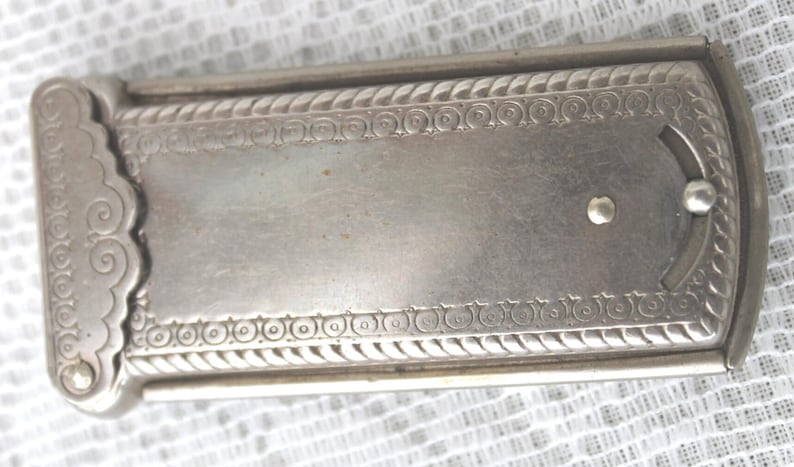 May include: A rectangular, silver-toned metal object with ornate detailing. The piece features a decorative border with a rope-like design and a scalloped edge on one side. A small, circular knob is visible.