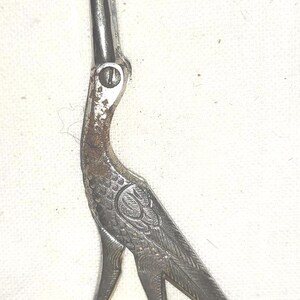 May include: Vintage silver-toned embroidery scissors in the shape of a bird. The scissors feature intricate detailing on the bird's body and head, with a curved beak and a detailed feather design. The blades are sharp and pointed.