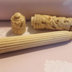 Puede incluir: Juego de escritura antiguo de color marfil, tallado intrincadamente, que incluye una pluma, un portalápices y un pequeño recipiente. La pluma tiene un diseño acanalado, mientras que el portalápices y el recipiente presentan tallas florales y arquitectónicas detalladas.