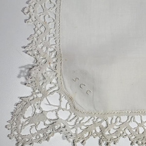 Op de afbeelding: Een witte zakdoek met een delicate, opengewerkte kanten rand. Het kant heeft een crèmekleurige off-white tint en de stof lijkt van zacht katoen of linnen te zijn. De initialen "SCC" zijn in de hoek geborduurd.