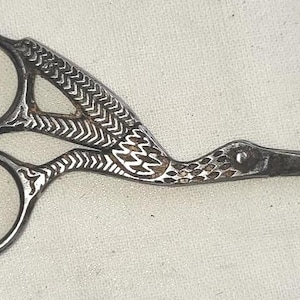 May include: Antique silver-toned embroidery scissors in the shape of a bird. The scissors feature detailed patterns on the handles and a pointed beak. The scissor's rings are circular, and the blades are sharp.