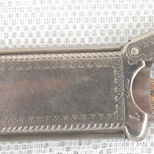 May include: A vintage silver-toned metal cigarette case with an ornate design. The case has a hinged lid with a decorative edge and a small latch. The interior reveals a comb-like structure. The number 7 is visible.