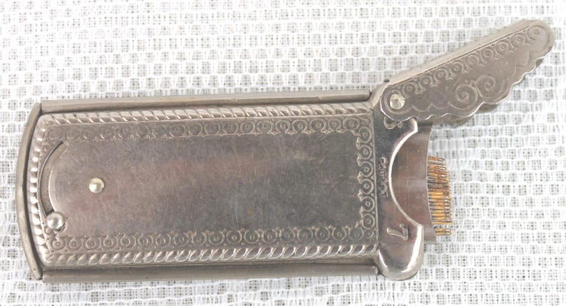 May include: A vintage silver-toned metal cigarette case with an ornate design. The case has a hinged lid with a decorative edge and a small latch. The interior reveals a comb-like structure. The number 7 is visible.