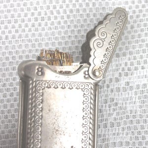 May include: A silver-toned needle case with an ornate design. The case is rectangular with a hinged lid, and the lid is open, revealing a bundle of needles. The number "8" is visible on the side.