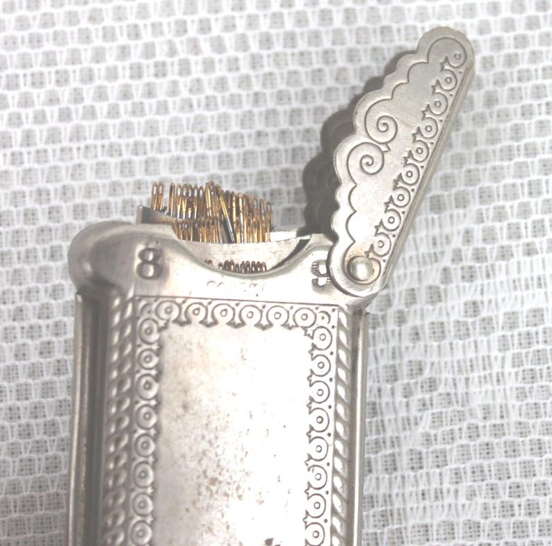 May include: A silver-toned needle case with an ornate design. The case is rectangular with a hinged lid, and the lid is open, revealing a bundle of needles. The number "8" is visible on the side.