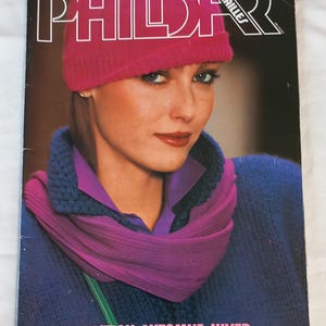 Può includere: Una copertina di rivista con una donna che indossa un cappello di lana rosa, una sciarpa viola e un maglione blu. Il titolo della rivista "PHILDER" è in grandi lettere bianche. La copertina include anche il testo "COLLECTION AUTOMNE-HIVER" e informazioni sui prezzi.