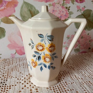 Puede incluir: Una tetera de cerámica blanca con un diseño floral con flores amarillas y azules. La tetera tiene una tapa y un asa.