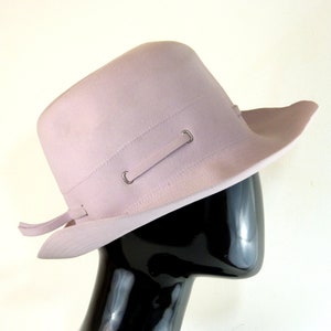 Op de afbeelding: Een lichtroze wollen fedora hoed met een brede rand en een band met metalen accenten.