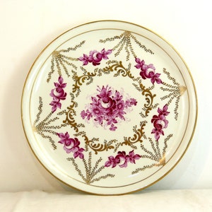 May include: A white ceramic plate with a gold rim and a floral design. The flowers are pink and white and are arranged in a circular pattern. The plate is decorated with gold accents.